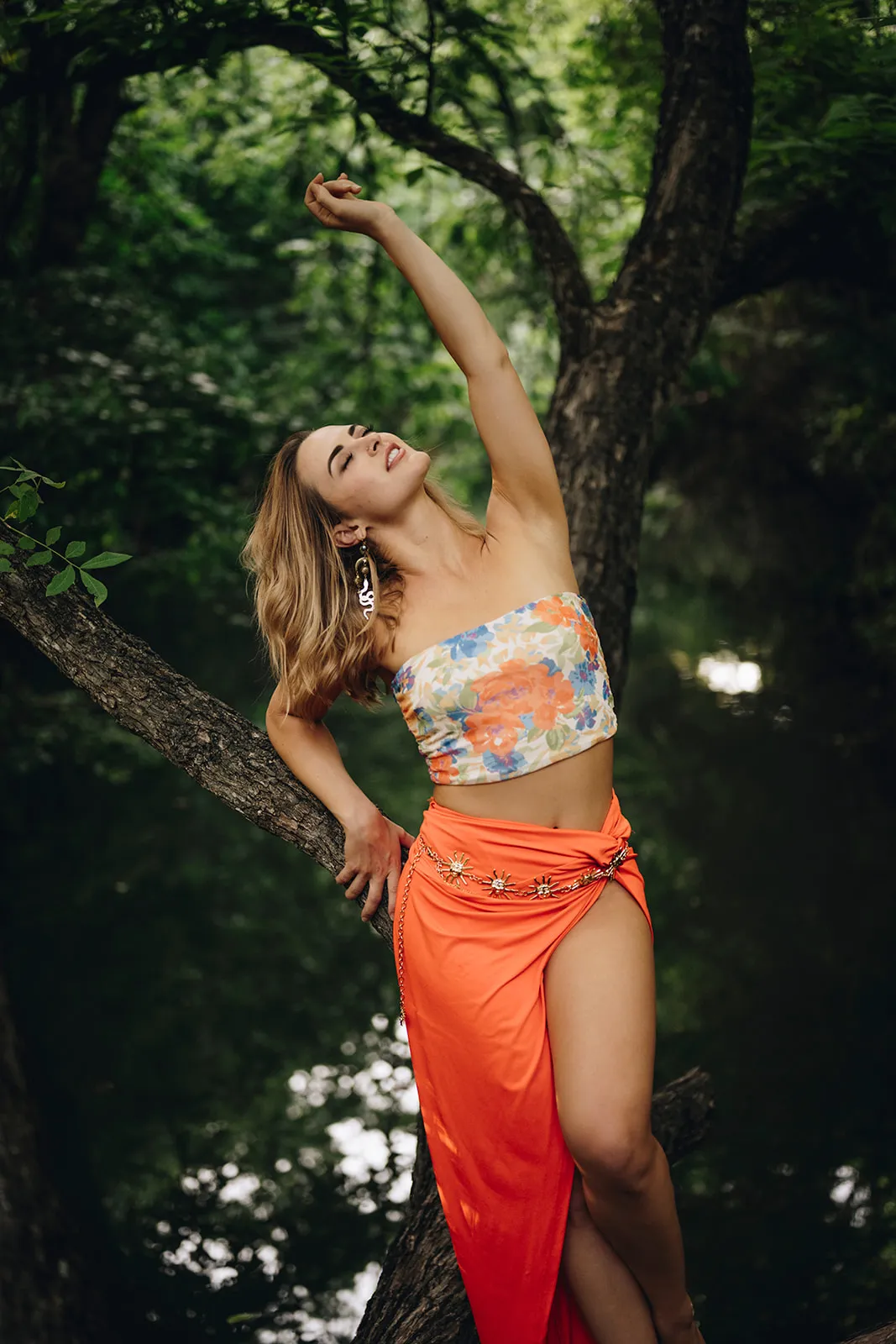 Taylor Tatum reaching up toward the trees in an orange skirt by the river